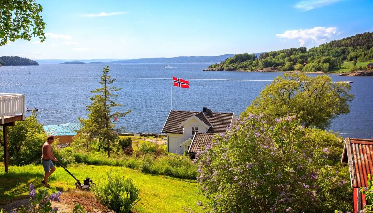 Man klipper gr&auml;set vid ett hus med norsk flagga vid en vacker fjord. En idyllisk sommardag i son Norge.