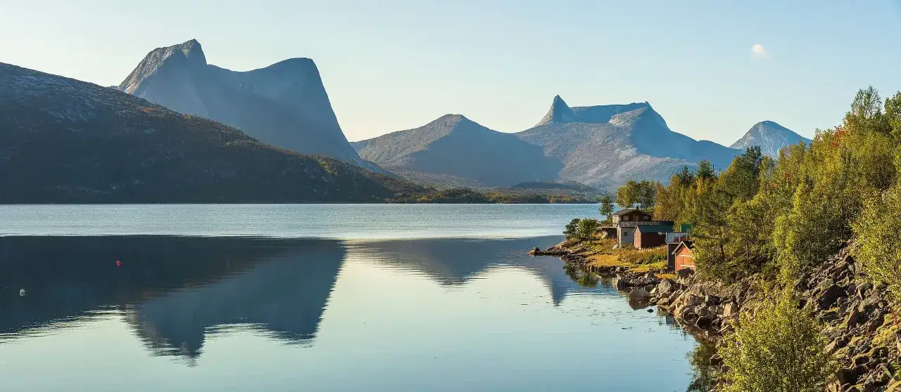 Majest&auml;tiska berg speglas i lugnt vatten vid en idyllisk kust med r&ouml;da hus. Perfekt f&ouml;r vandring i Narvik.