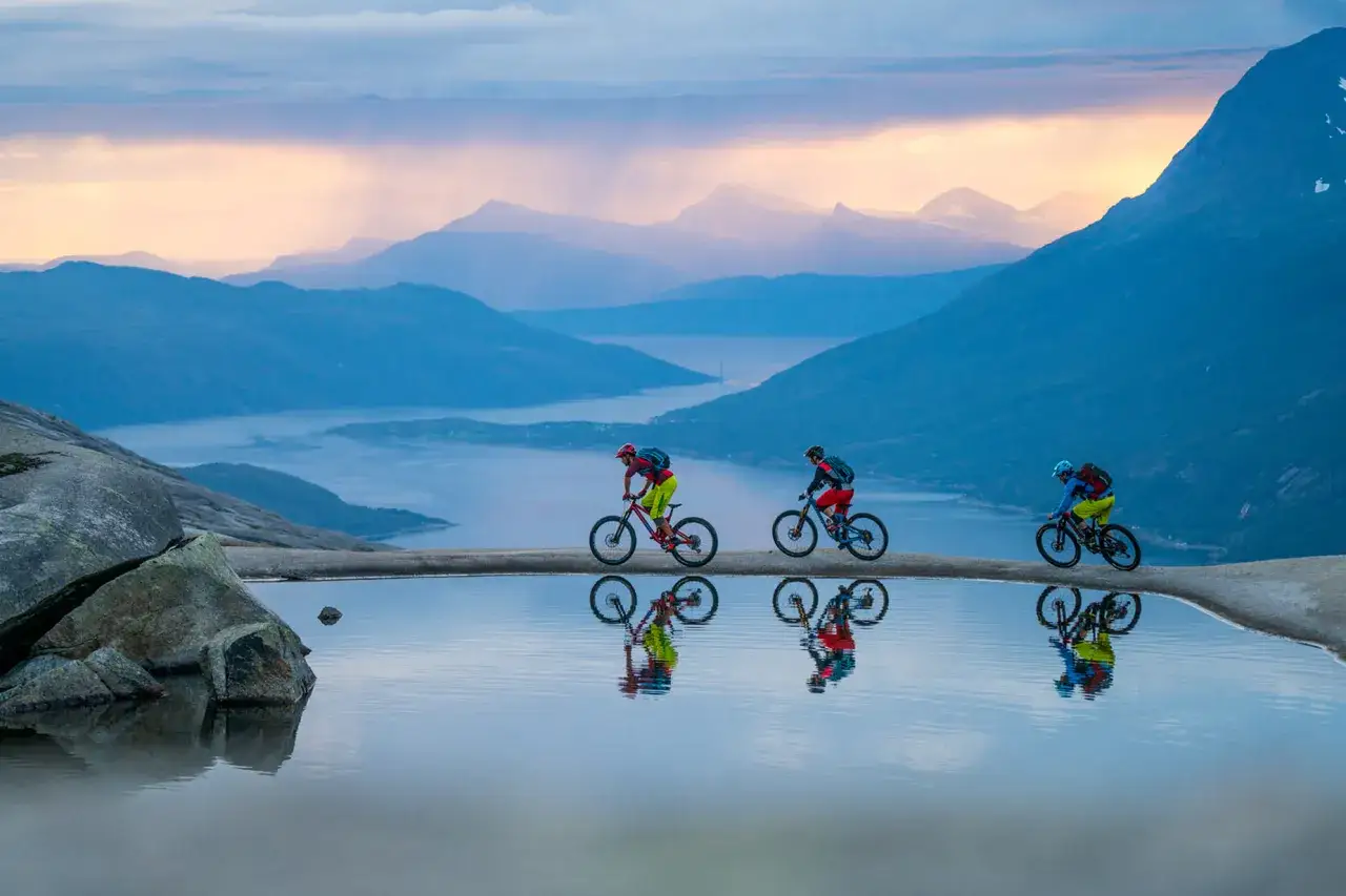 Tre cyklister på mtb norge susar fram längs en klippkant med en spegelblank vattenpöl. Dramatiska berg och en fjord i bakgrunden.