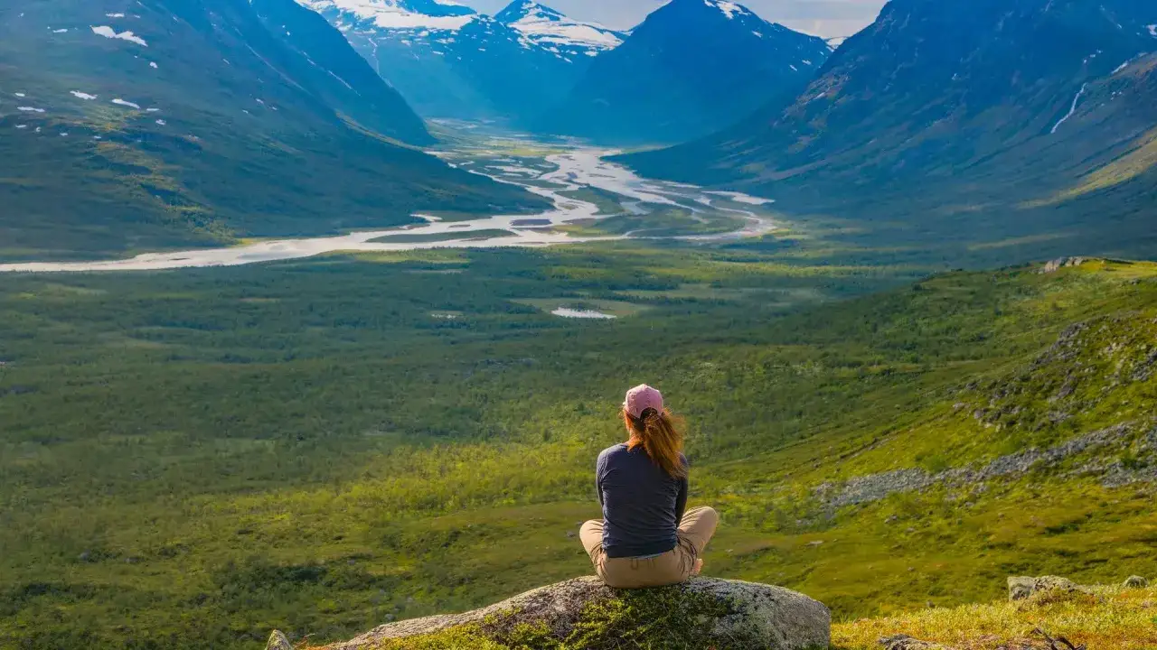 En person sitter på en sten och blickar ut över en vidsträckt dalgång i Sarek nationalpark. Snöklädda berg reser sig i bakgrunden.