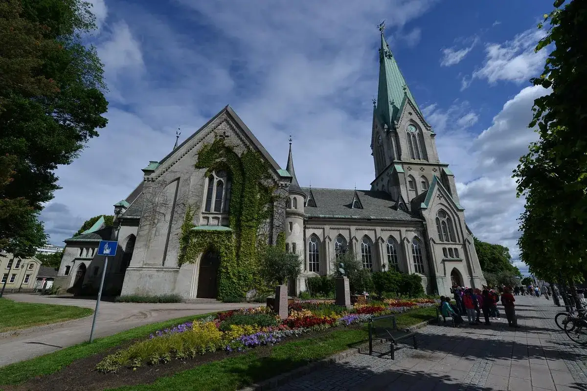 Imponerande kyrka med torn och gr&ouml;nt tak i Kristiansand, Norge. Blommor och tr&auml;d pryder omgivningen.