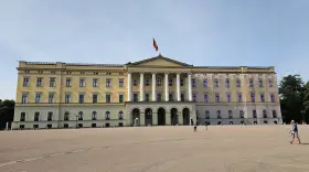 Det gula **Oslo slott** med sin flagga reser sig stolt mot en klarblå himmel. En stor öppen plats framför slottet.