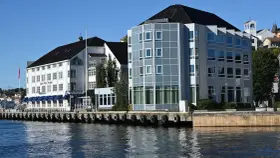 Hotellbygg vid vattnet i Arendal, Norge. En vit byggnad med blå markiser och en modern glasbyggnad står sida vid sida.