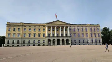 Det gula **Oslo slott** med sin flagga reser sig stolt mot en klarblå himmel. En stor öppen plats framför slottet.