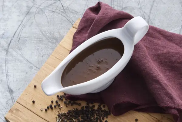 En vit såsslev fylld med mustig brunsås, bredvid en hög med svartpepparkorn. Ett perfekt brunsås recept för middagen.