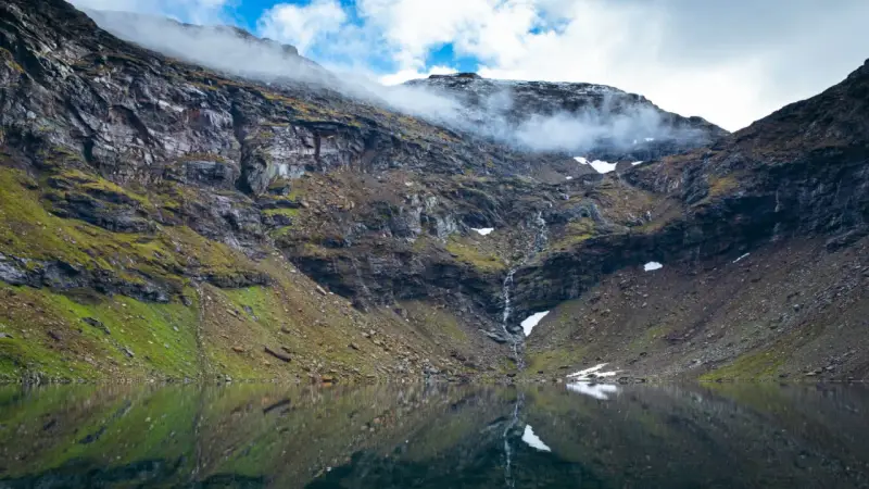 En spegelblank sjö reflekterar en dramatisk bergssida med en forsande vattenfall. Perfekt för att vandra i Abisko.