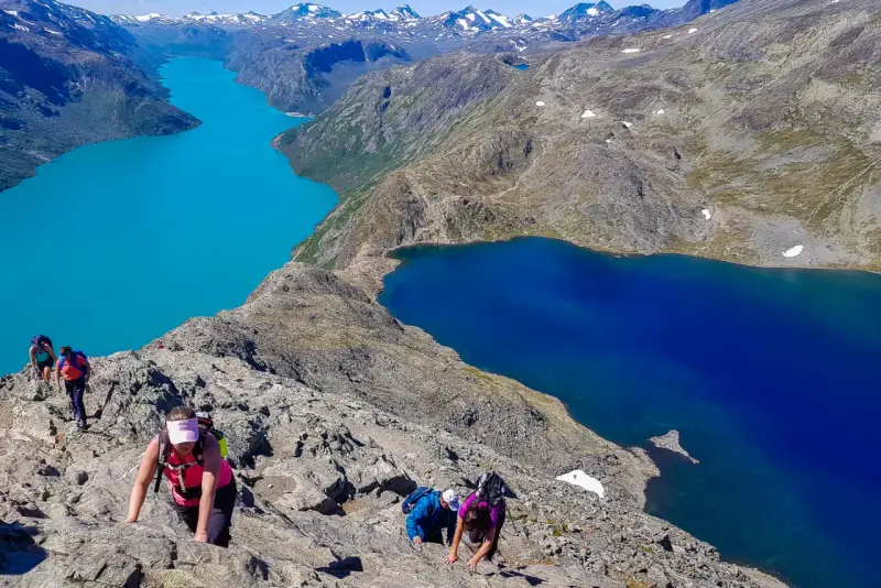 Vandrare klättrar på en stenig bergssida med turkosa och blå sjöar nedanför. En klassisk Besseggen vandring.