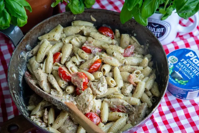 En stor panna med krämig pasta pesto, körsbärstomater och kyckling. En träsked ligger i pannan.