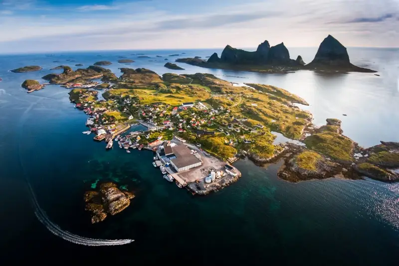 En pittoresk by på en ö i Norge, med berg i bakgrunden. En båt lämnar ett spår i vattnet. En vacker vy över traena norge.